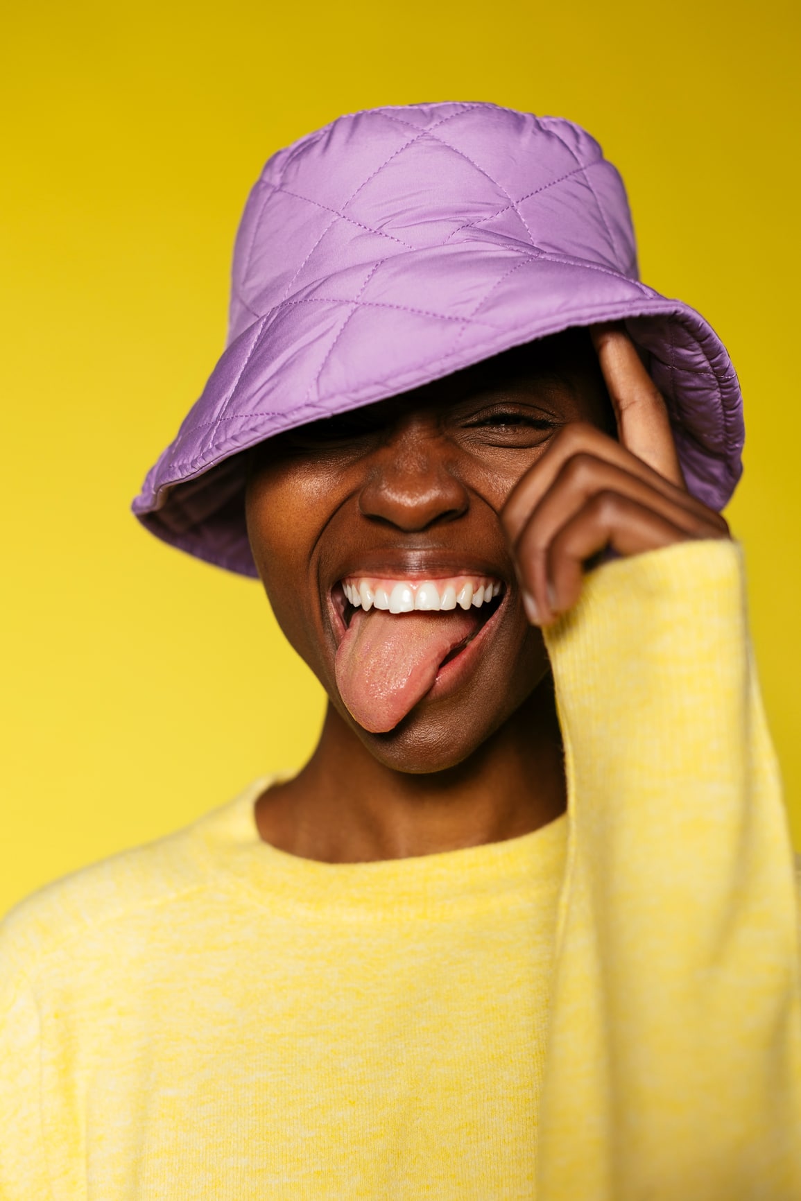 Cheerful female wearing yellow sweater and trendy purple hat showing tongue in yellow background in studio