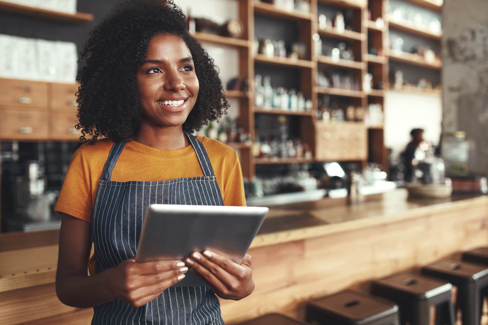 Close-up of a young female owner holding digital tablet in hand smiling and daydreaming in a coffee shop