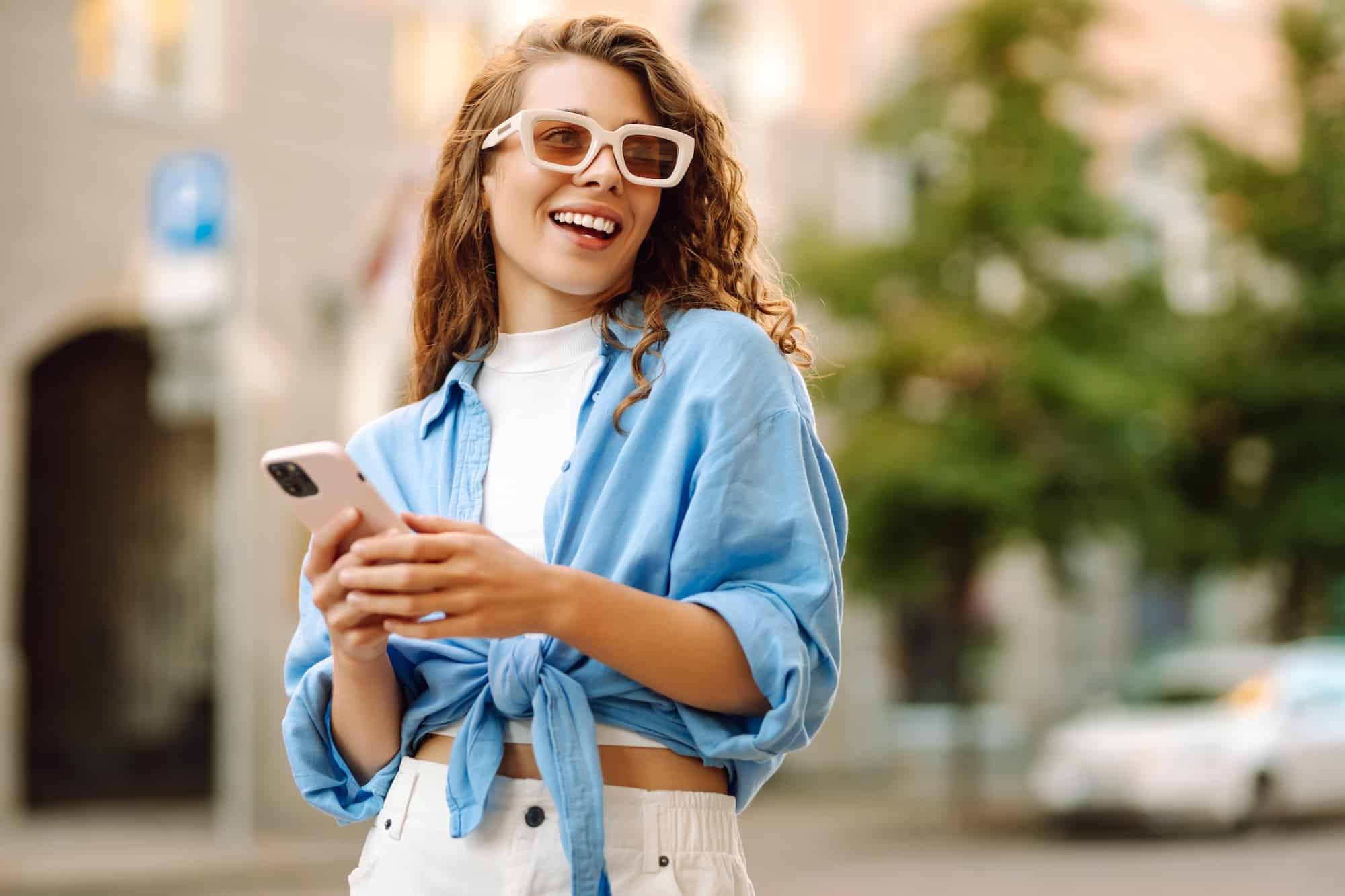 Cheerful young woman in fashionable sunglasses engages with her smartphone.