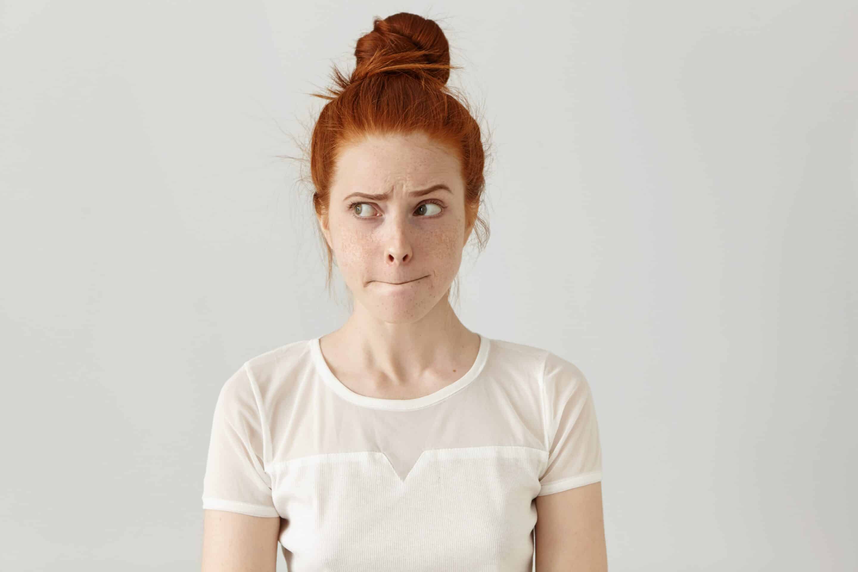 Indoor shot of cute redhead girl looking away, having doubtful and indecisive face expression, pursuing her lips as if forbidden to say anything. Confused young female posing isolated at white wall